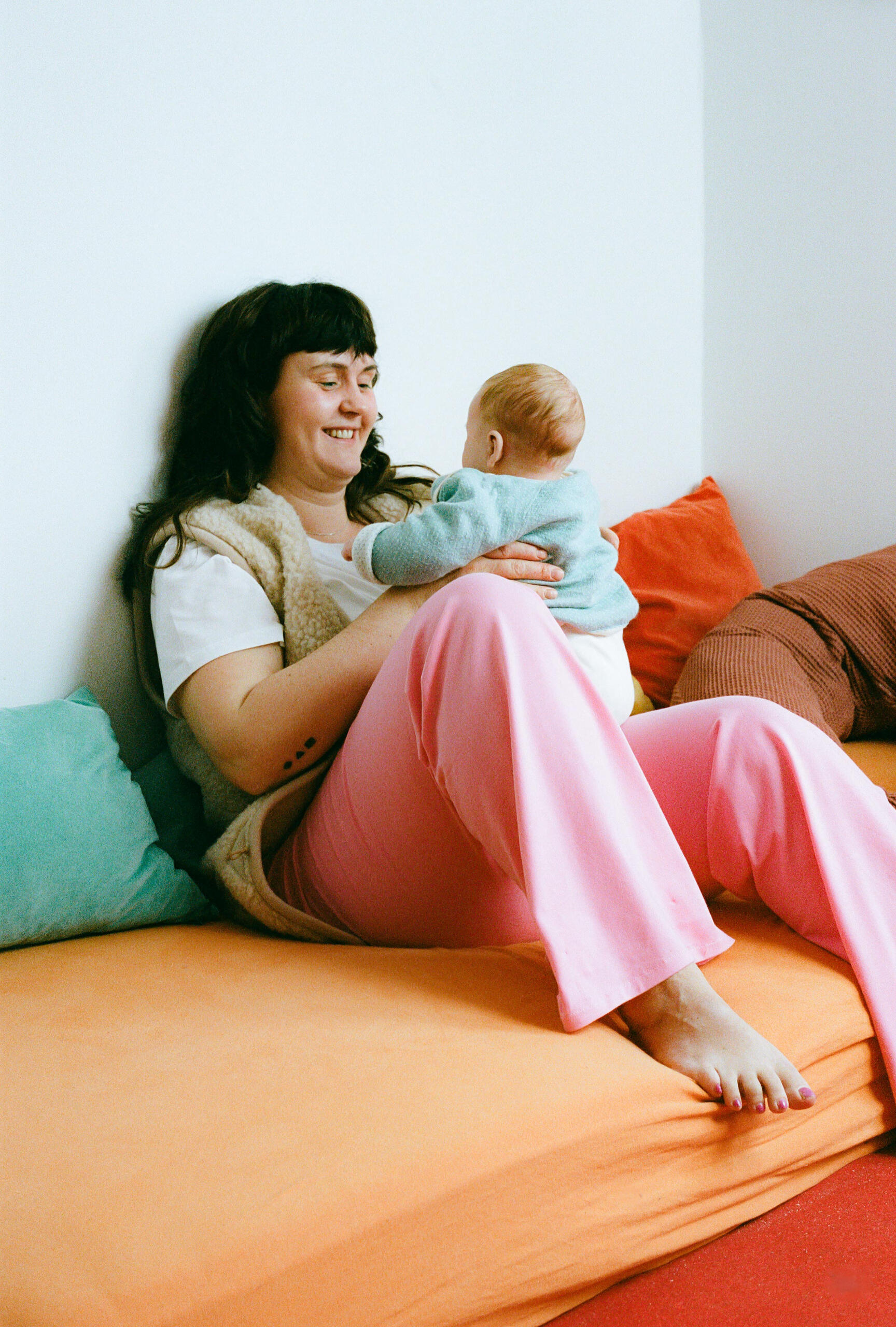 Greta schaut ihr Baby auf ihrem Schoß an Mutter und Baby teilen einen liebevollen Blick, während die Mutter MIUMI’s Umstandshose mit weitem Bein trägt.