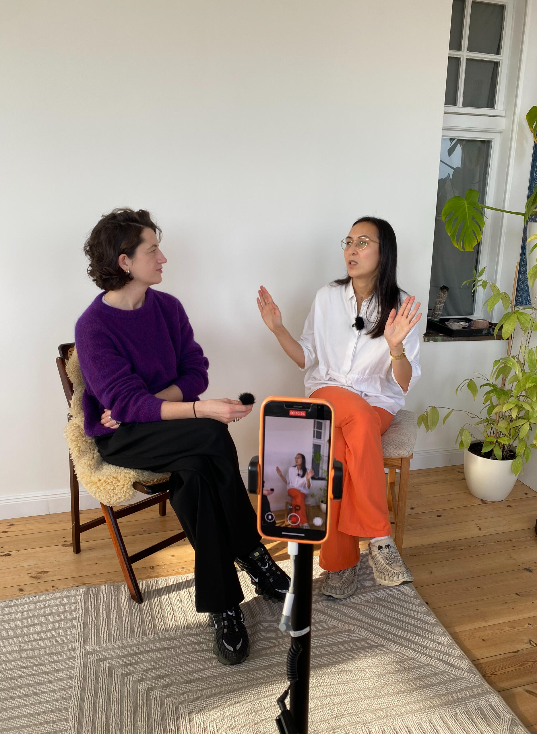 Breathwork teacher Kim Gerlach in a MIUMI interview with Caecilia. Two women are sitting in a bright room in the middle of an interview; one is wearing a purple sweater, the other a white top and orange MIUMI pants. A smartphone on a tripod records the conversation.