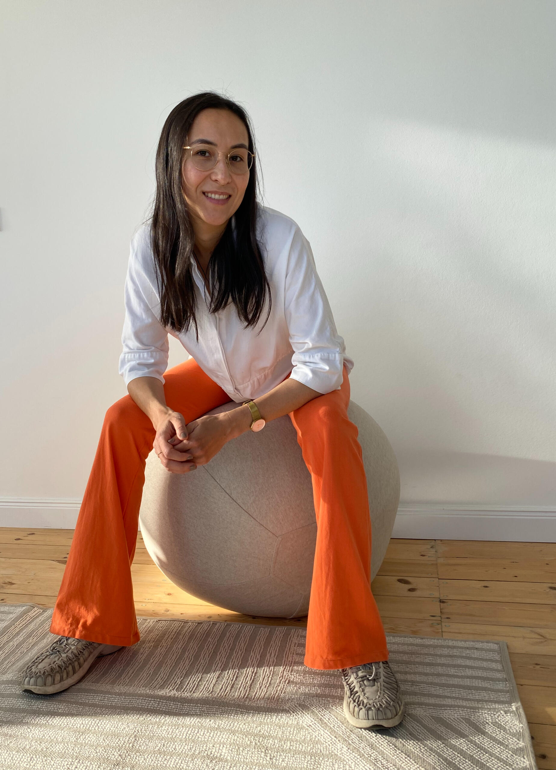 Breathwork Lehrerin Kim Gerlach in einer orangen MIUMI Schlaghose. Eine Frau mit langen dunklen Haaren und Brille lächelt, während sie auf einem grauen Gymnastikball sitzt. Sie trägt ein weißes Hemd, eine orangefarbene Schlaghose von MIUMI und beigefarbene Schuhe. Der Schauplatz ist ein heller Raum mit Holzboden und einer