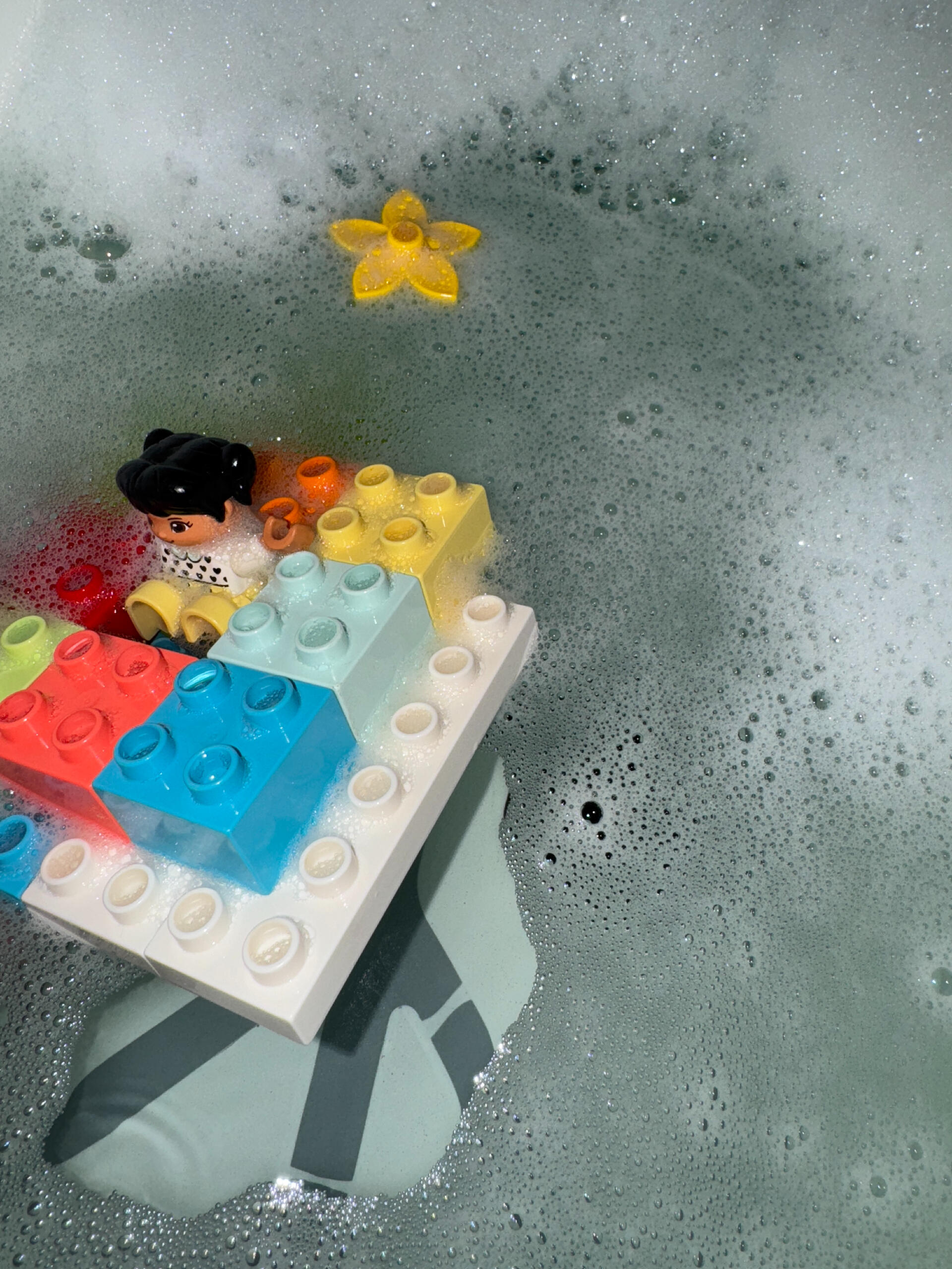 LEGO boat A toy figure sits on a boat made of colorful LEGO bricks amidst soapy bubbles in a bath. A yellow star-shaped toy floats nearby.