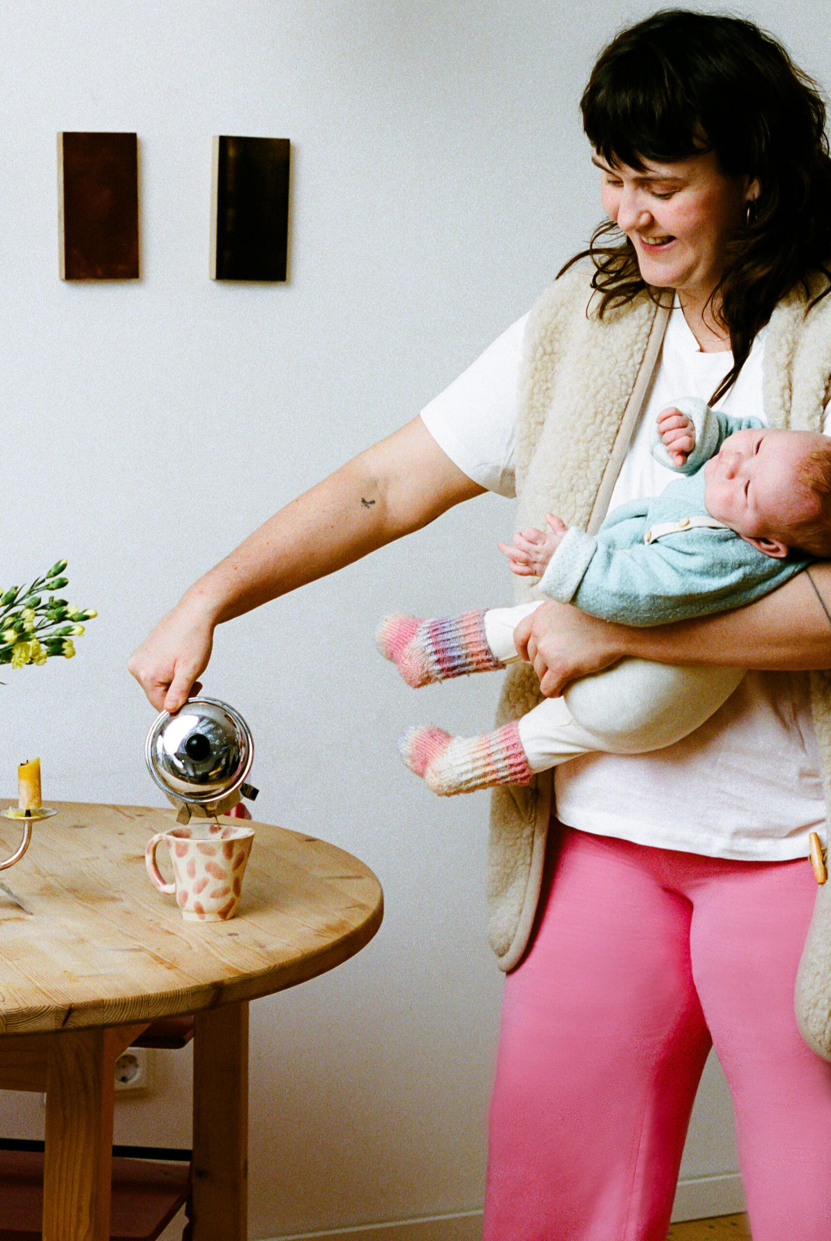 Greta poring herself a coffee Mother in MIUMI’s pink sustainable maternity pants, holding her baby and enjoying a coffee in the kitchen.
