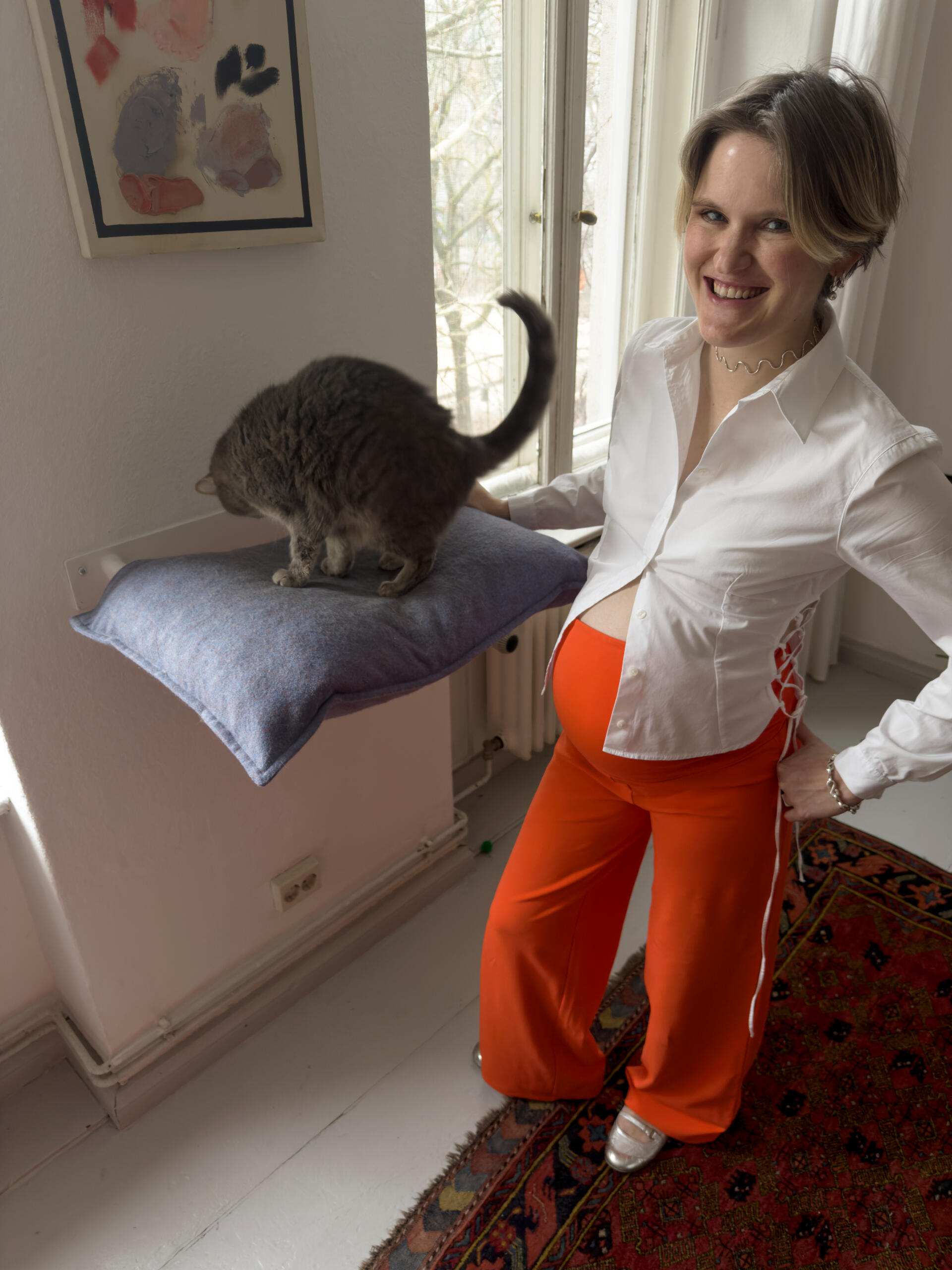Sonja in MIUMI wide leg pants with her cat at home. Smiling woman with baby belly in a white blouse and vibrant orange MIUMI maternity pants. Next to her sits a gray cat on a pillow. Bright room with abstract art and large windows. Warm and joyful mood.