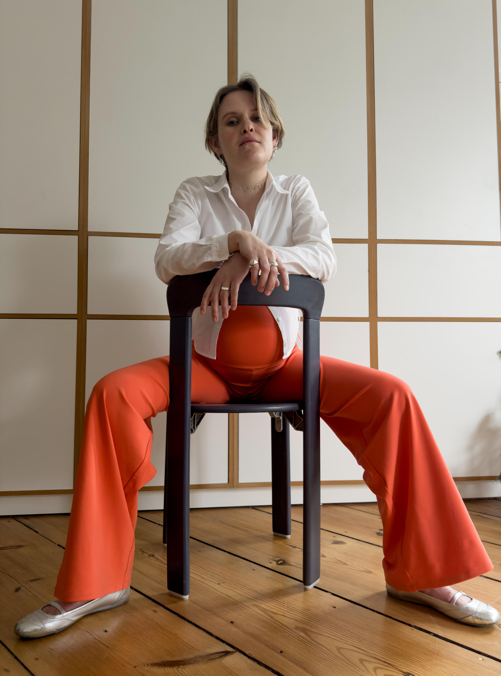 Sonja in MIUMI Wide Leg Pants in orange. Schwangere Frau in weißer Bluse und leuchtend orangefarbener MIUMI-Umstandshose sitzt rückwärts auf einem Stuhl.
