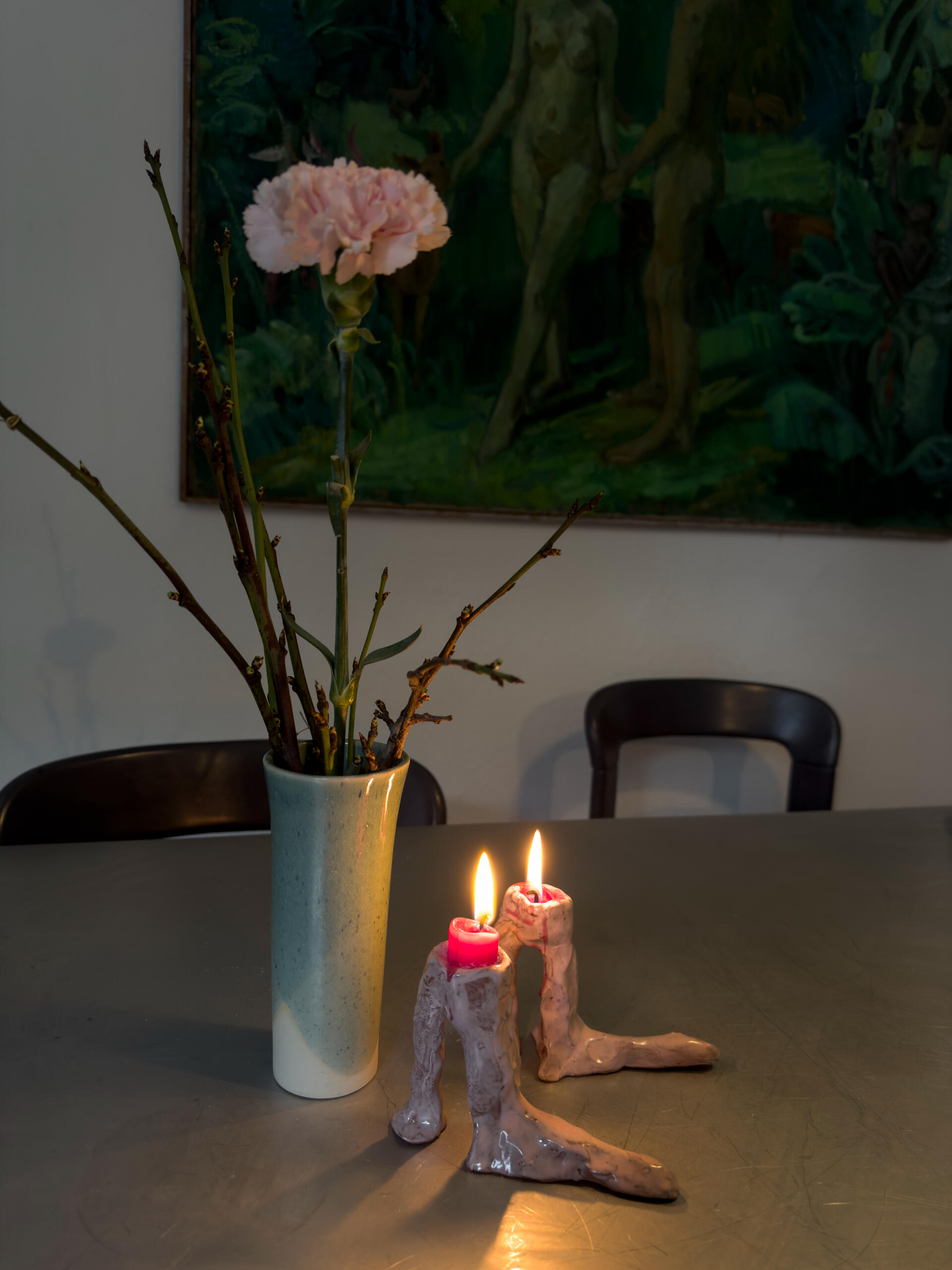 Detail aus Sonjas gemütlichem Zuhause. Eine rosa Nelke in einer Vase, umgeben von kahlen Zweigen, steht auf einem Tisch neben zwei brennenden Kerzen in einem skurrilen Kerzenhalter. Im Hintergrund hängt ein grünes Gemälde.