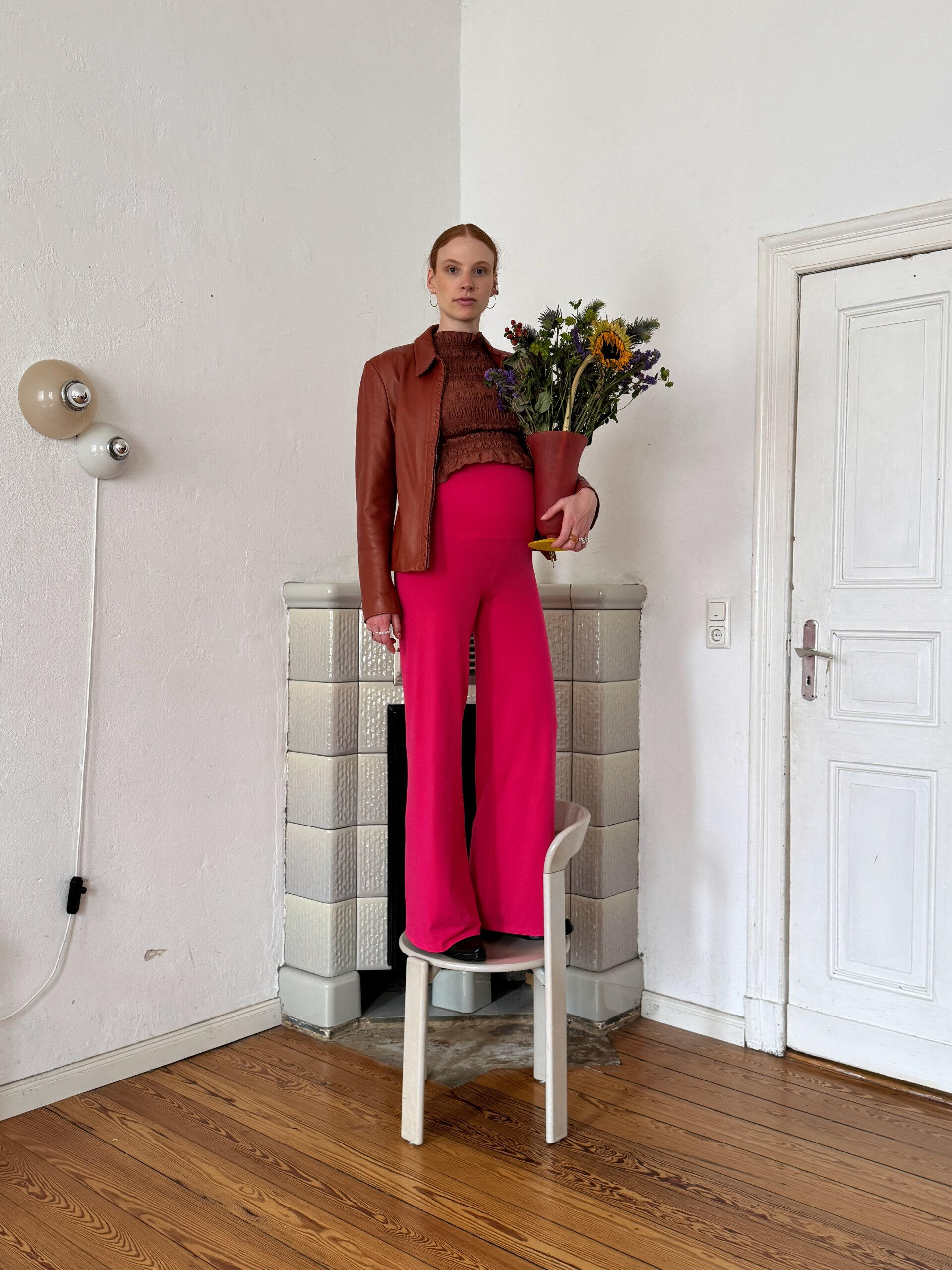 Katharina Bellinger liebt boldes Design Eine schwangere Frau in einer braunen Jacke und einer leuchtend pinkfarbenen MIUMI-Hose steht selbstbewusst auf einem Stuhl und hält eine Vase voller Trockenblumen vor einem minimalistischen Innenraum.