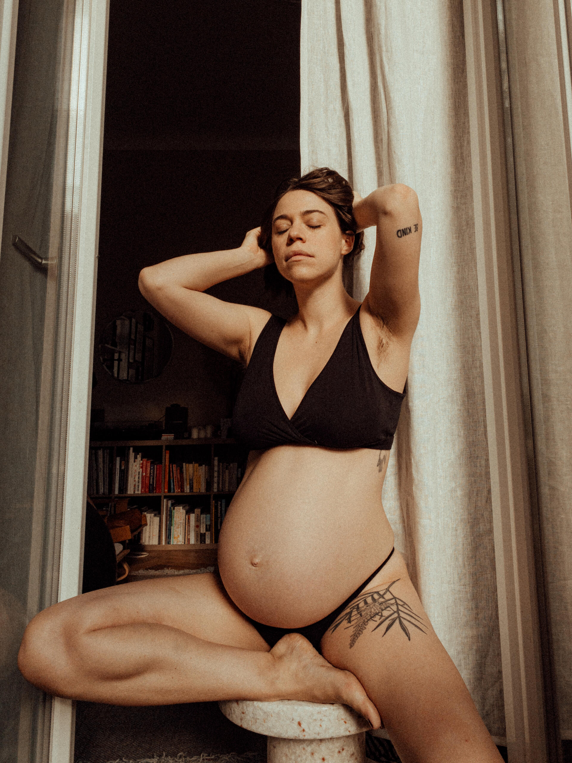 Eine schwangere Frau, nur in Unterwäsche, sitzt auf einem Stuhl vor einem weißen Vorhang. Ihre Augen sind geschlossen – ein intimer Moment, eingefangen im warmen Licht.