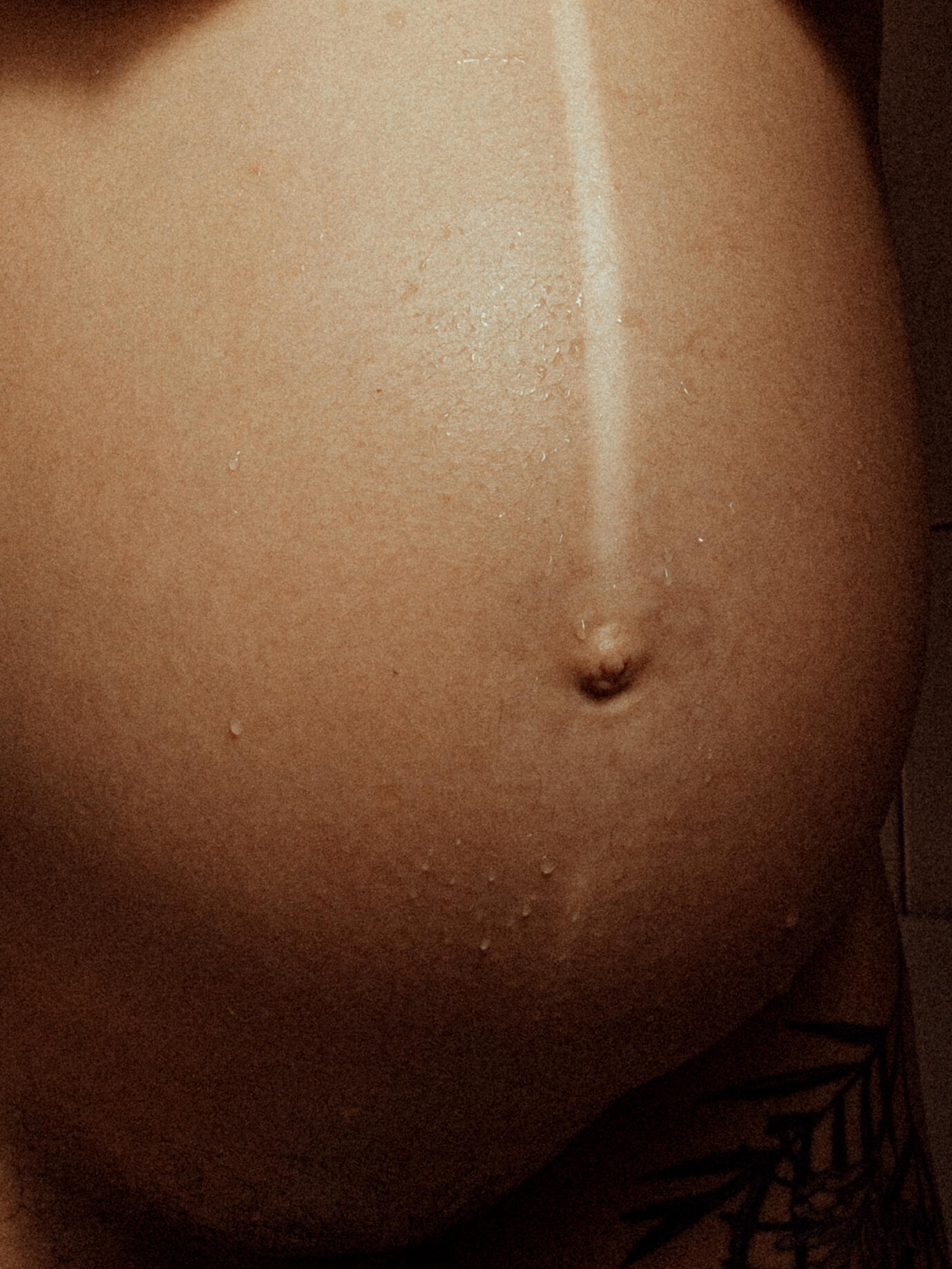 Nahaufnahme eines schwangeren Bauchs mit deutlicher Linea Nigra und Wassertropfen. Ein Tattoo blitzt unten rechts hervor – ein Bild voller Intimität und Stärke.