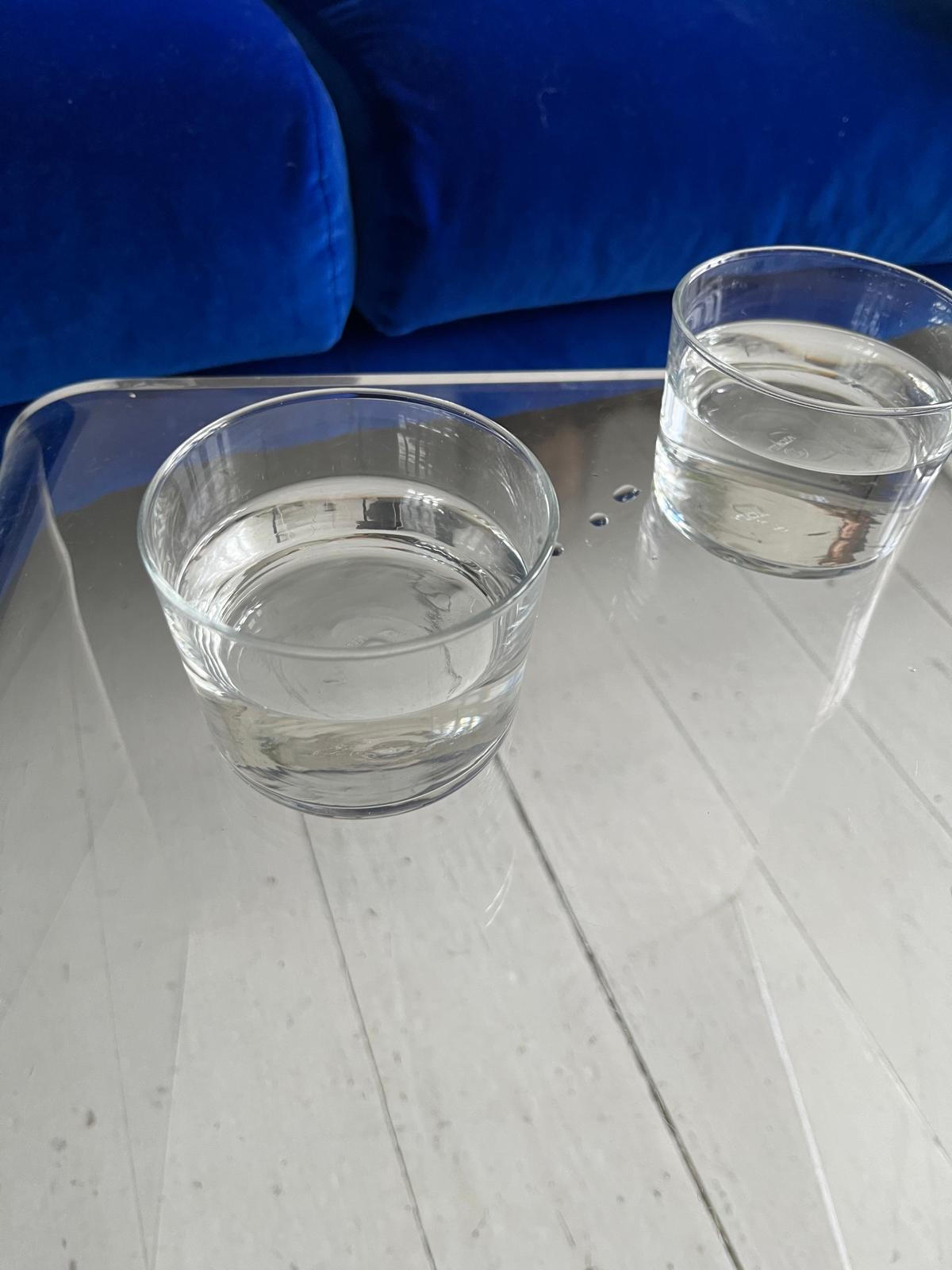 Two water glasses on a plexiglass table, with a blue velvet couch in the background. The setting is calm and modern.