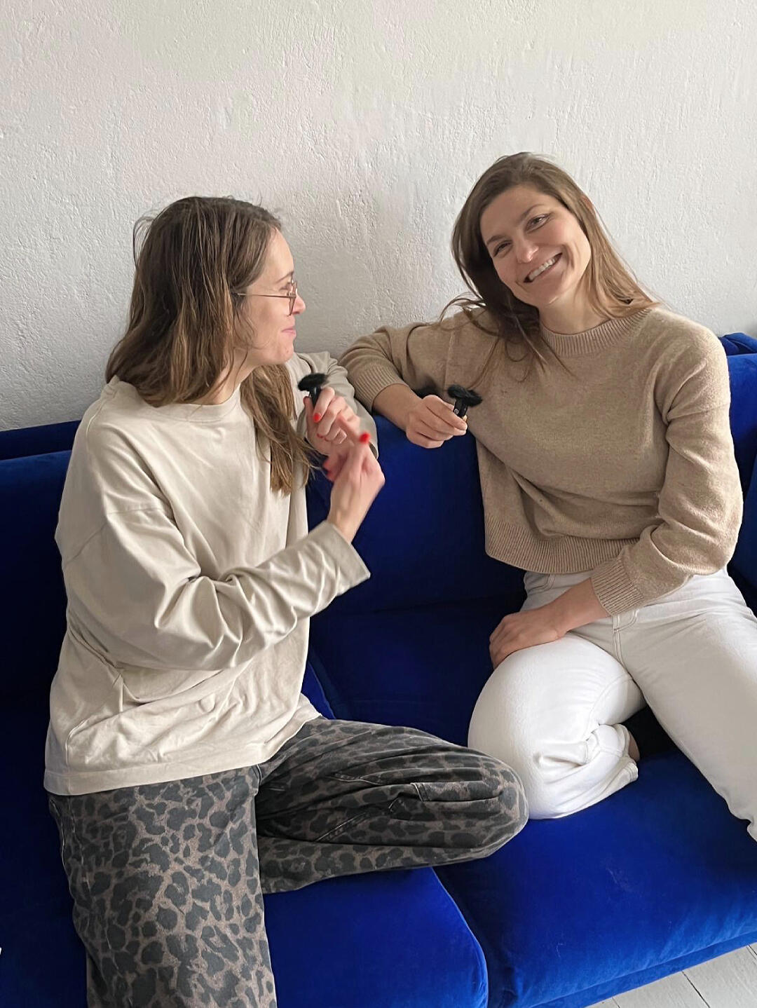 Zwei Frauen (Antonia Pahlke von MIUMI & Hanna Stutzki) sitzen auf einem blauen Sofa, lächeln und halten schwarze Klipp-Mikrofone in der Hand. Die eine trägt einen beigen Pullover, die andere ein cremefarbenes Oberteil und eine gemusterte Hose.